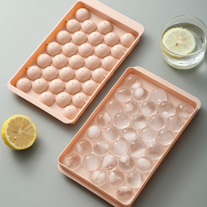 Two pink ice cube trays with filled ice cubes and water, lemons, and a glass on a gray surface.